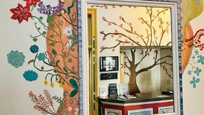 A wall with a mirror reflecting a room containing a desk, framed pictures, and a tree painting. - The Sheldon B&B (Eastbourne)
