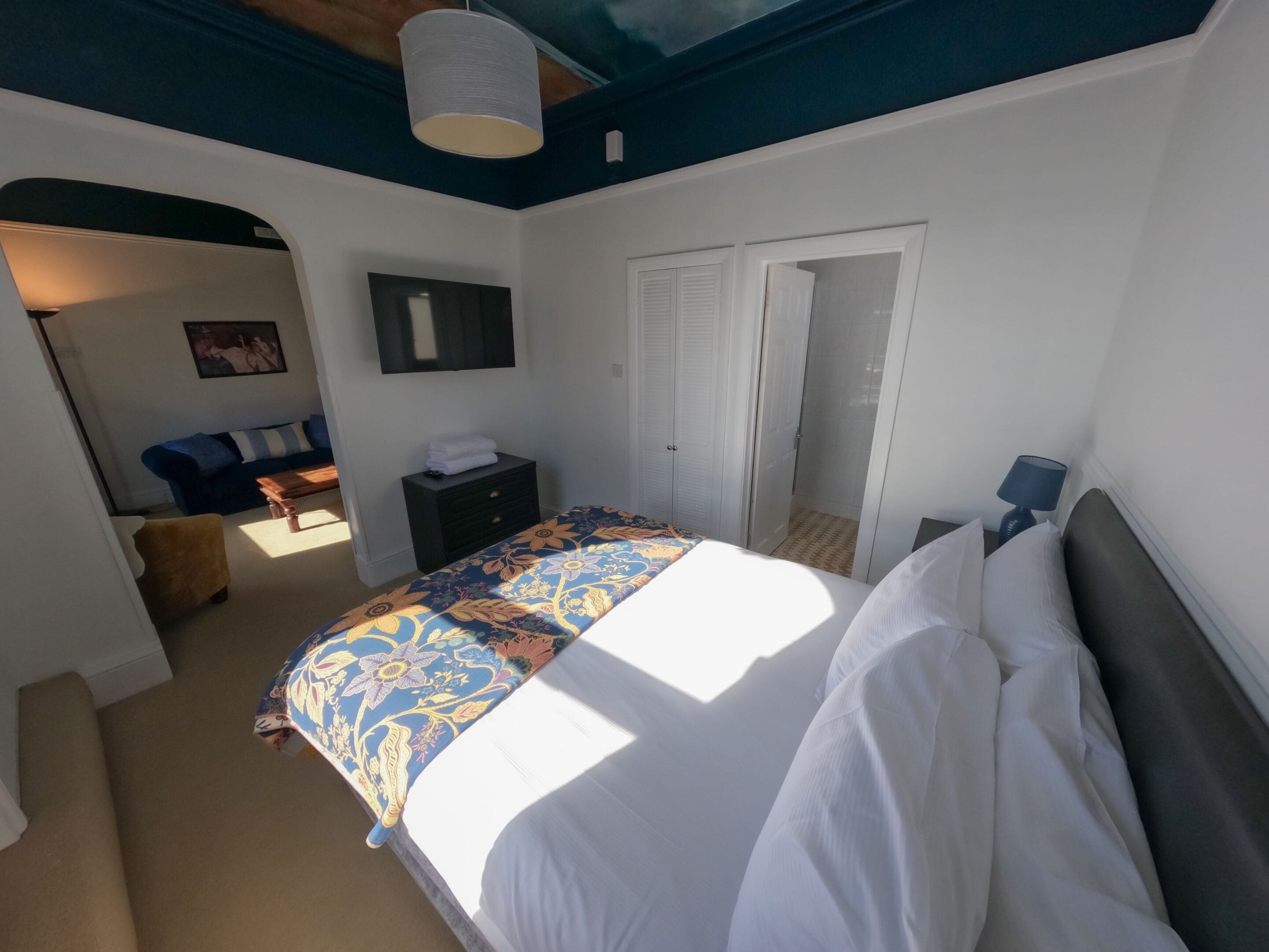 A bedroom with a bed, a dresser, a nightstand, a lamp, and a door leading to a bathroom.