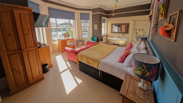 A bedroom with a large bed, a wooden wardrobe, a desk with a chair, a vanity with a mirror, and a window with a view of buildings. - The Sheldon B&B (Eastbourne)