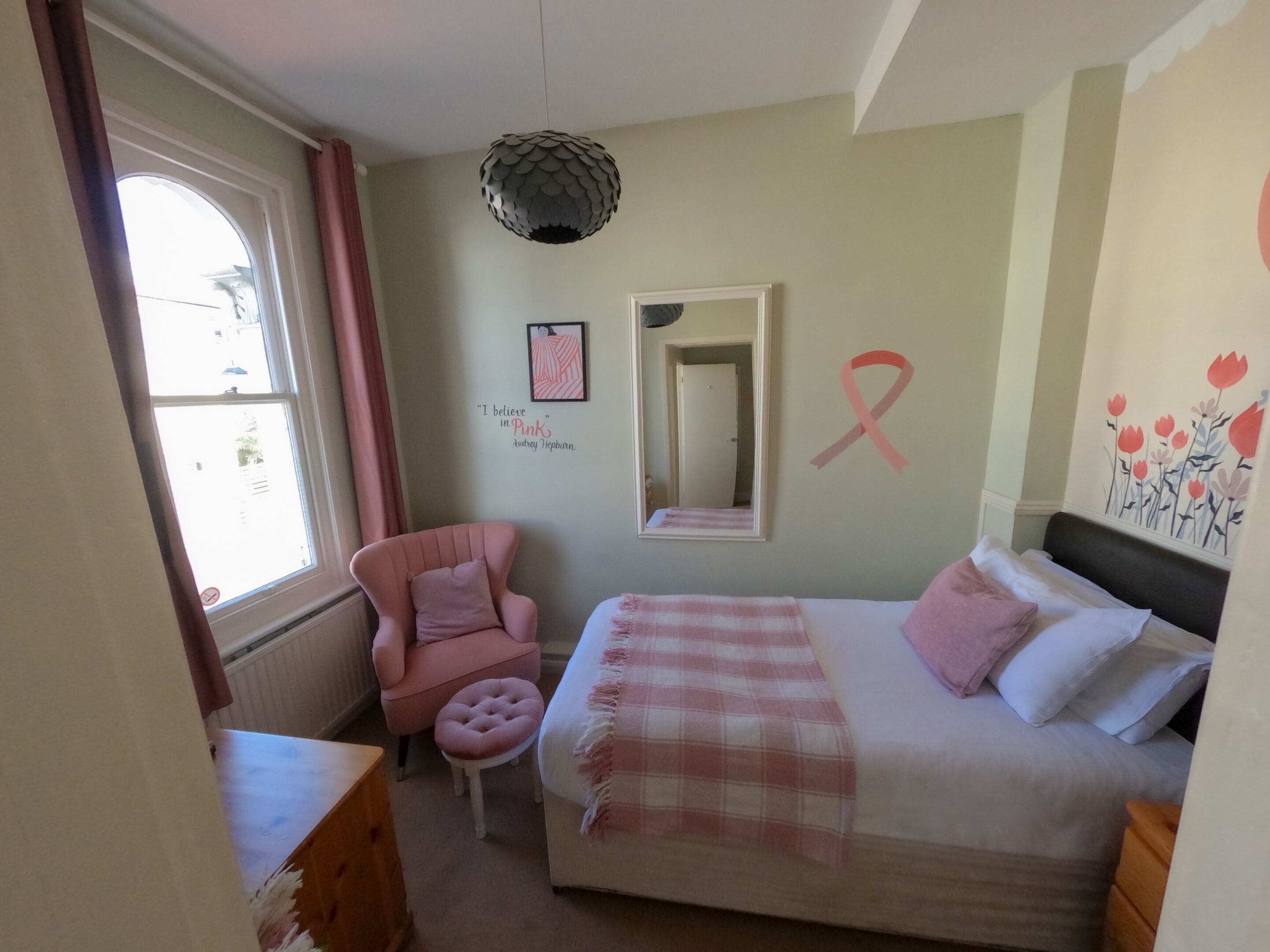 A bedroom with a bed, a chair, a mirror, a window, and a wall with a pink ribbon symbol.
