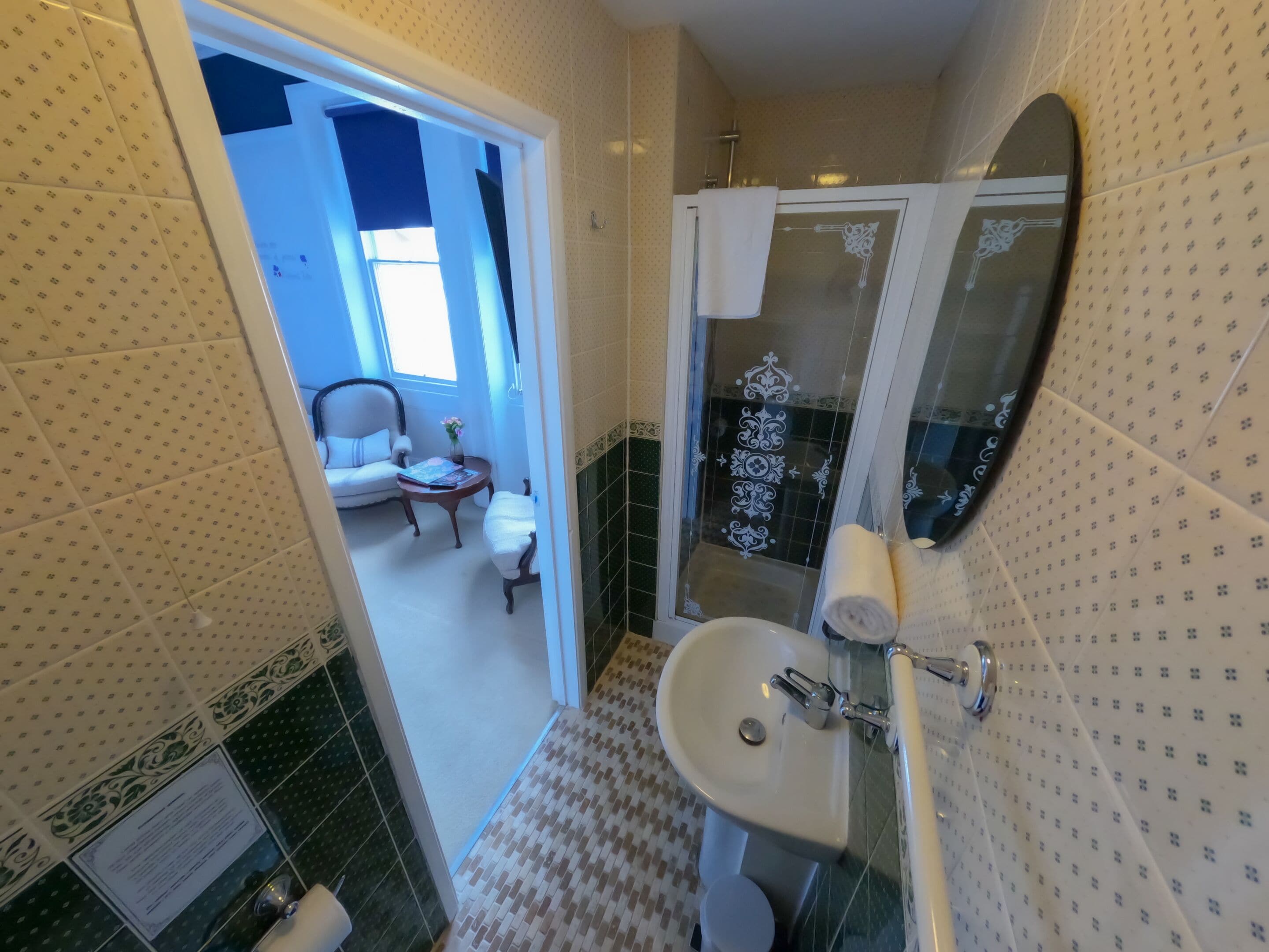A bathroom with a sink, mirror, and shower area. There is a chair and a small table with a vase in the adjacent room.