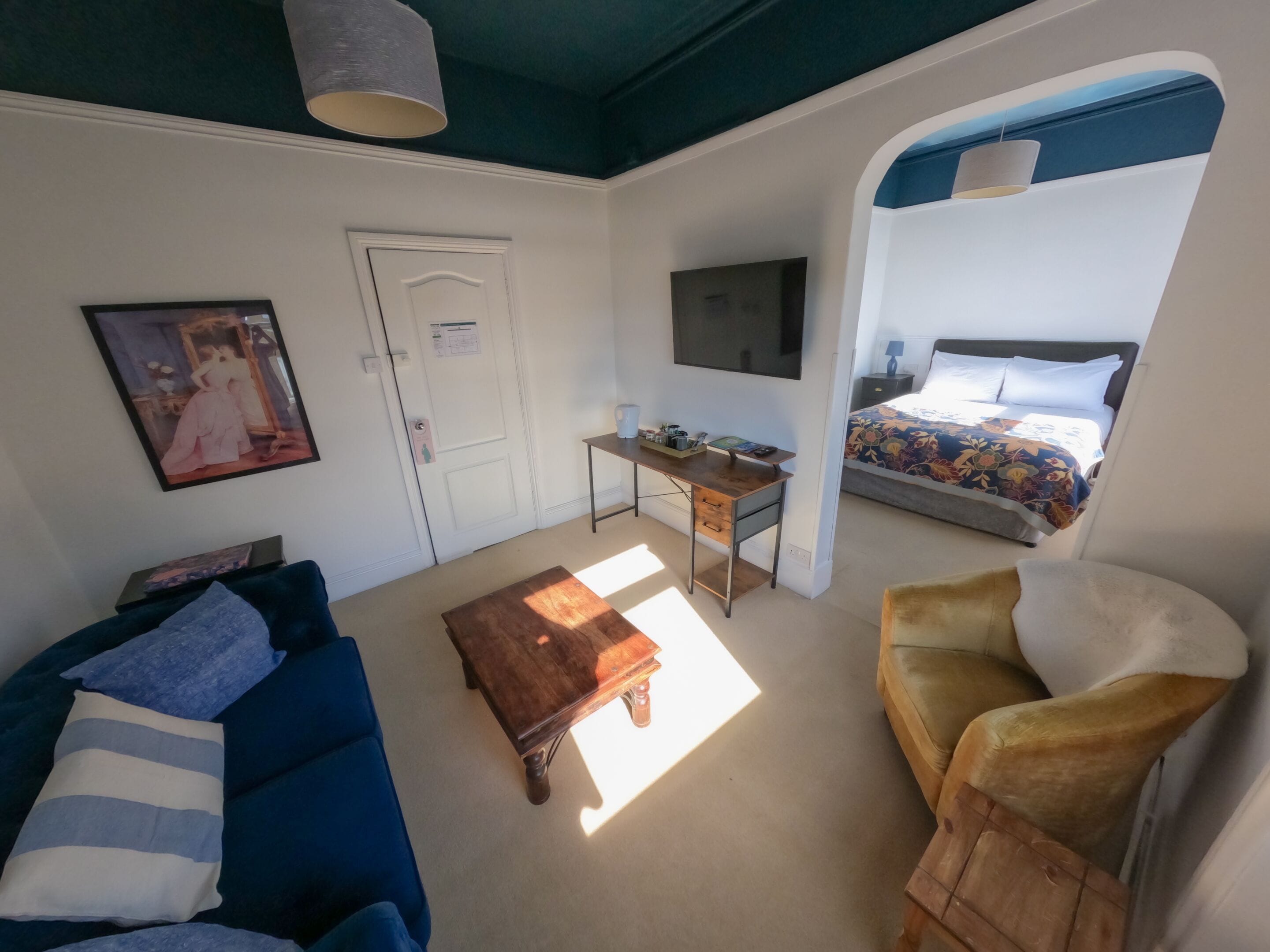 Quarto com cama, escrivaninha, sofá, poltrona, televisão e porta.