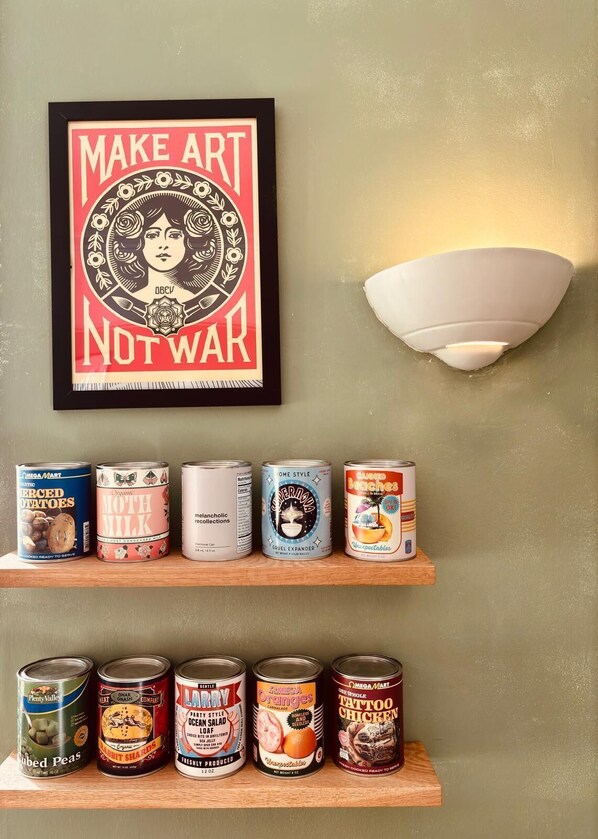 Una pared con un cartel enmarcado que dice "MAKE ART NOT WAR" y un aplique. Abajo, hay repisas con diversos productos enlatados.