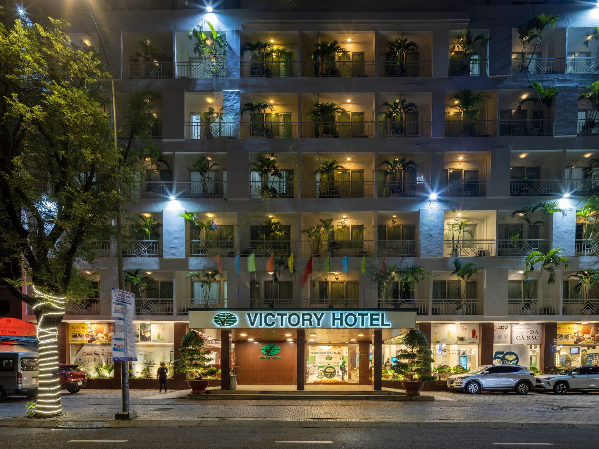 A multi-story hotel with balconies, a sign reading 'VICTORY HOTEL,' and a street view with cars and a tree.