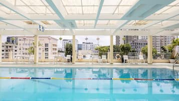 An indoor swimming pool with clear lane dividers, surrounded by white chairs and tables, with a view of buildings in the background.