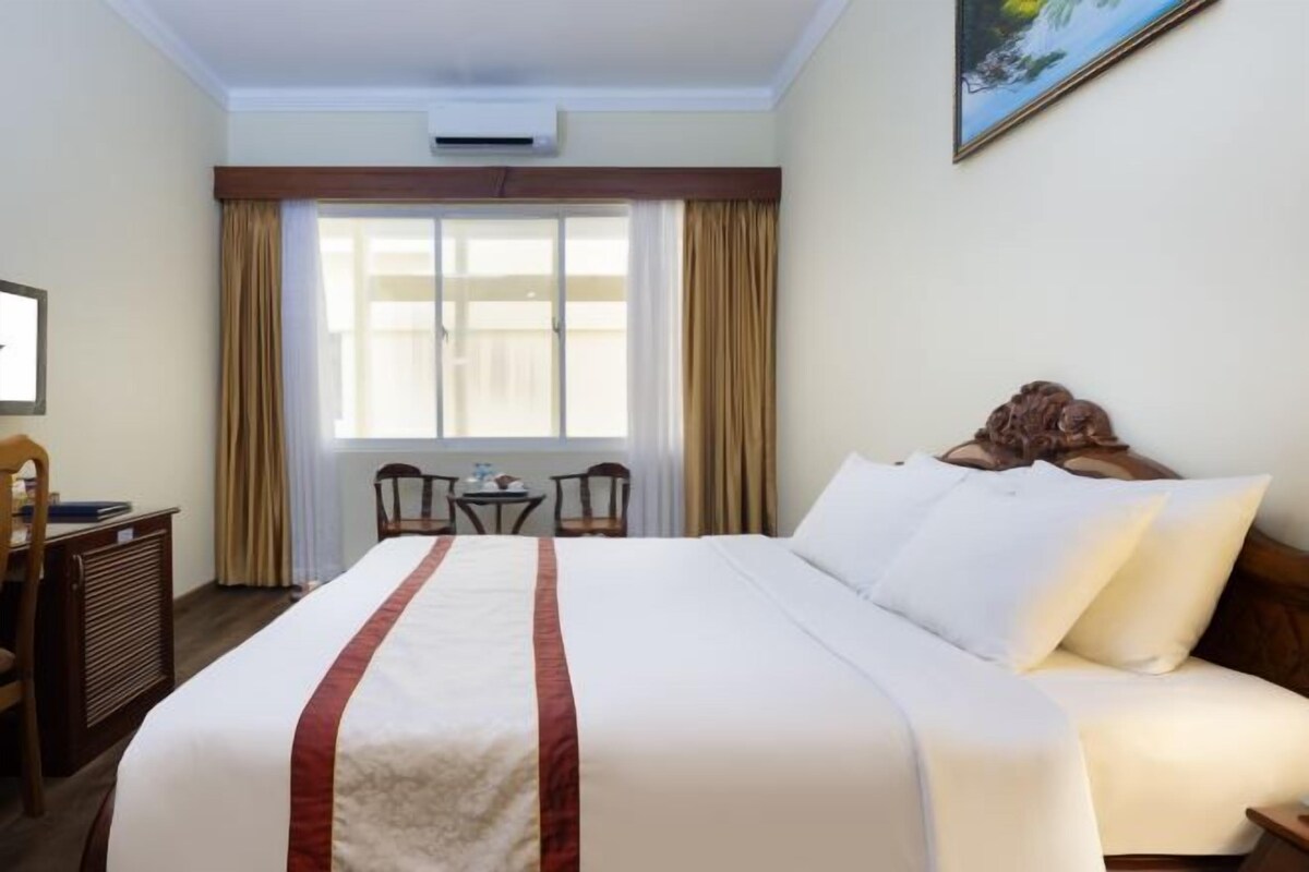 Quarto de hotel com uma cama grande, escrivaninha, cadeira e janela com cortinas.