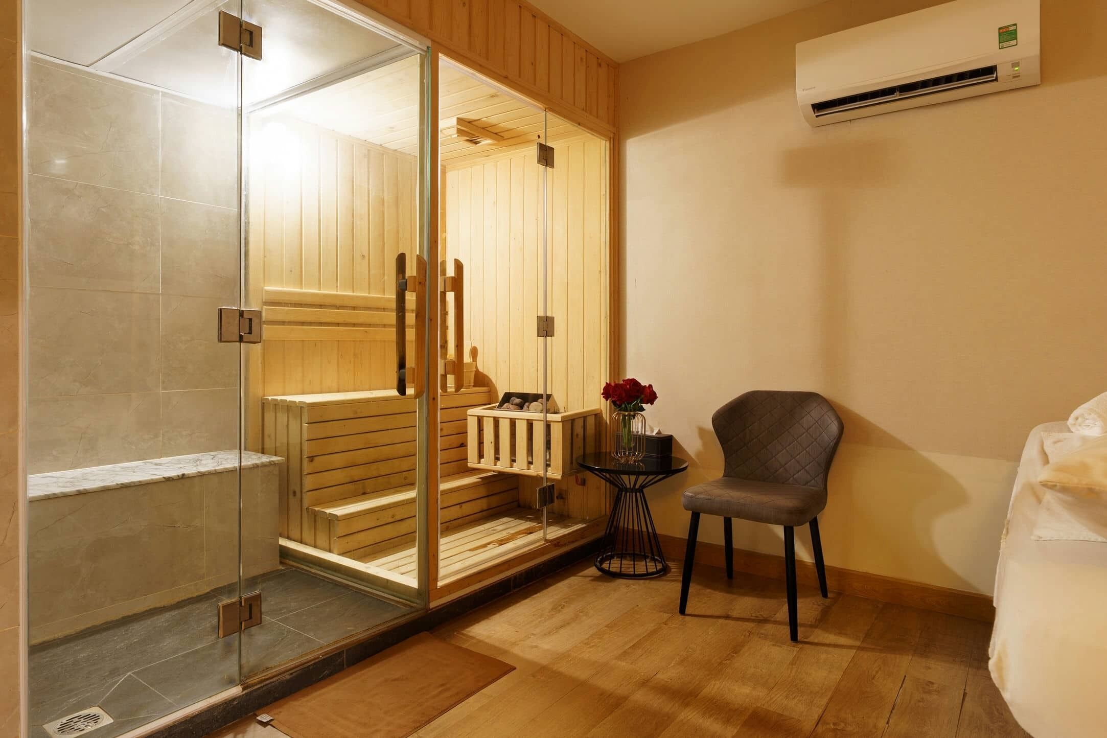 A sauna room with a glass door, wooden benches, a small table with a vase, and a chair.