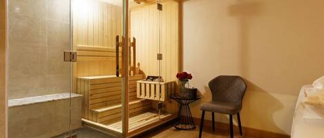 A sauna room with a glass door, wooden benches, a small table with a vase, and a chair.