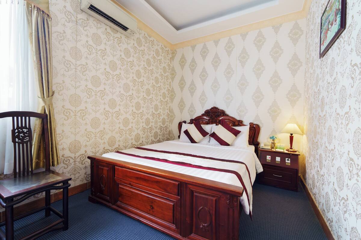 A bedroom with a wooden bed, a nightstand, a lamp, a chair, and patterned wallpaper.