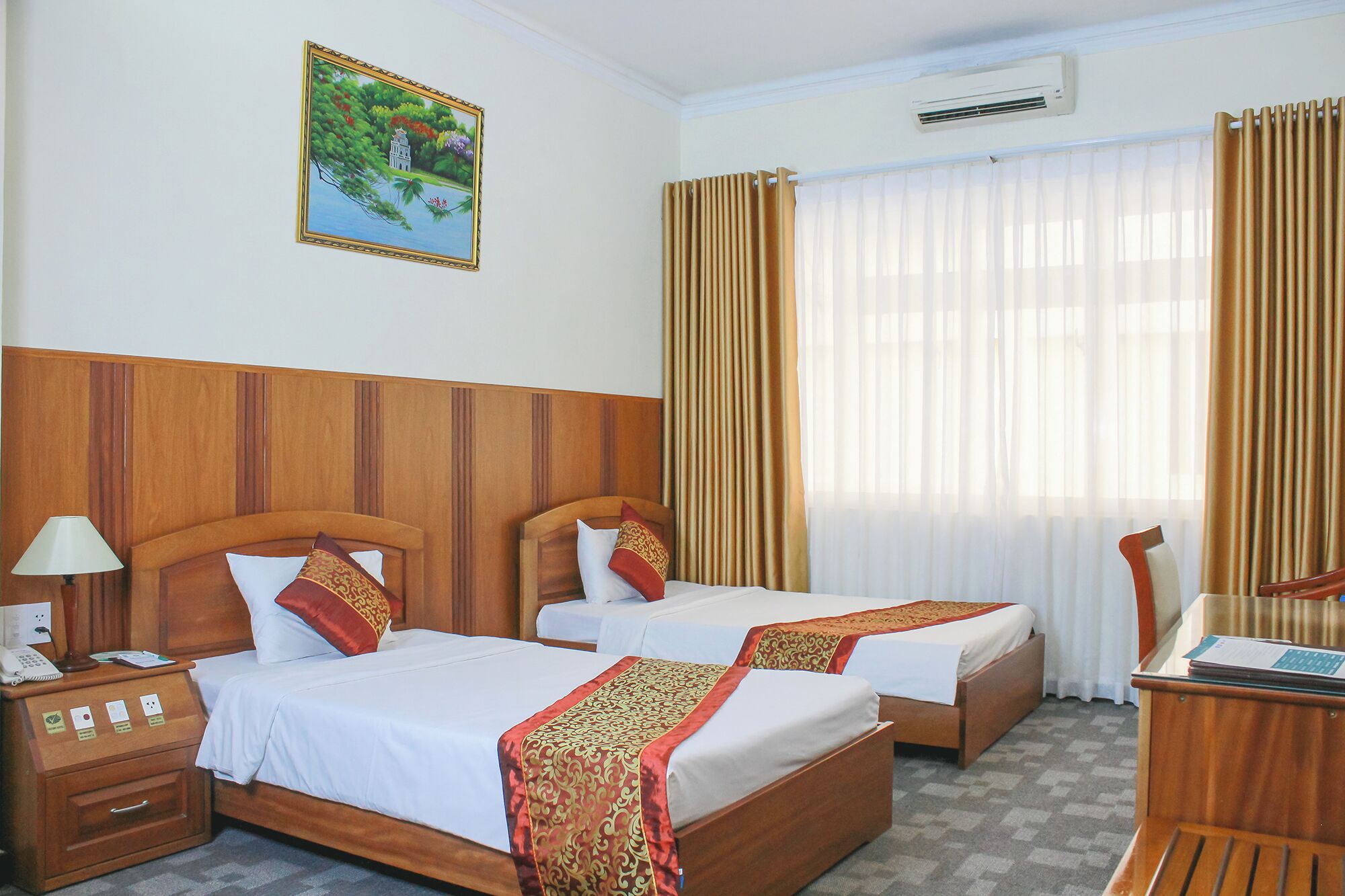 A hotel room with two beds, a wooden headboard, a nightstand with a lamp, a desk with a chair, and a painting on the wall.