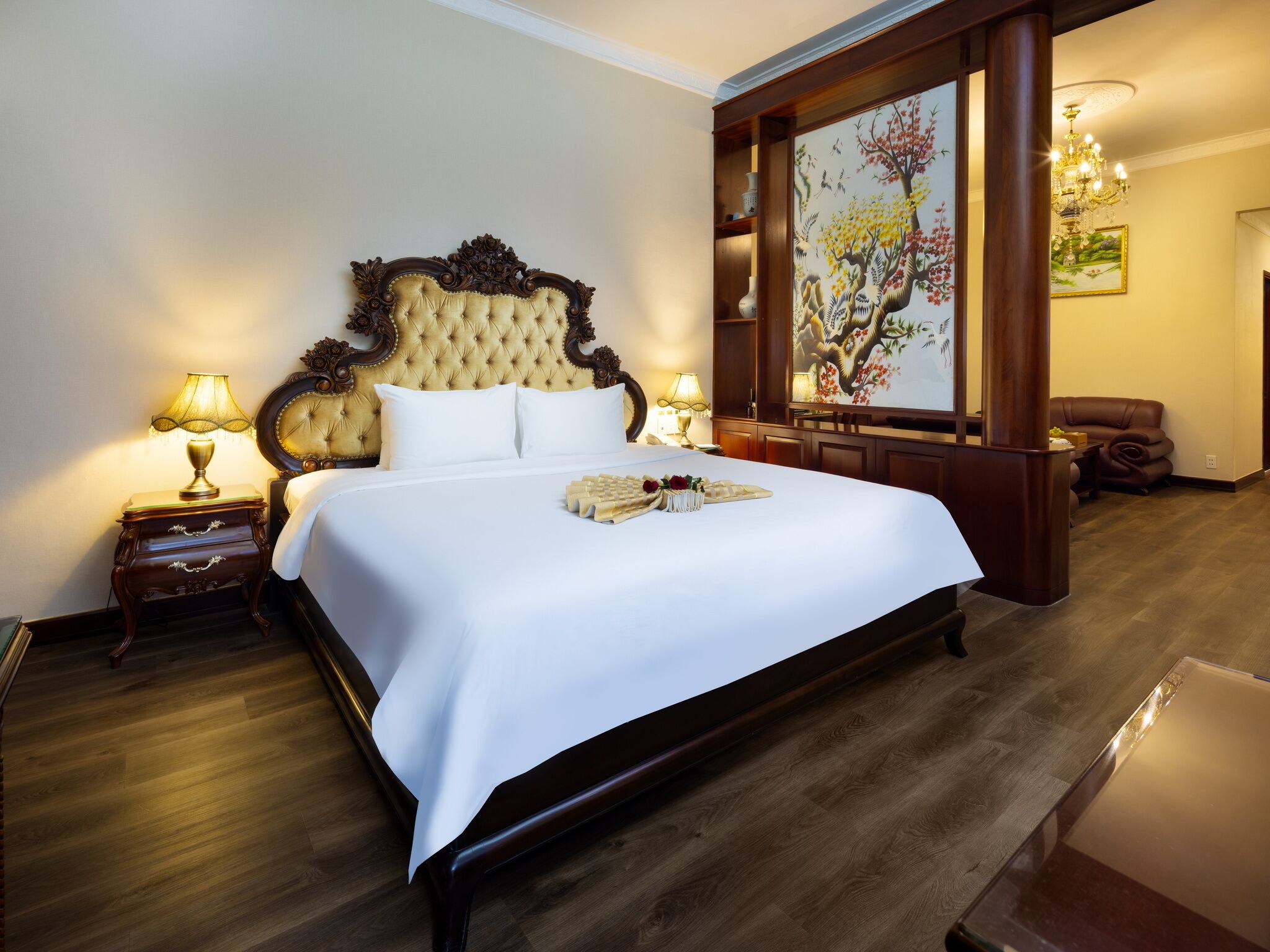 A large bed with a tufted headboard, two bedside tables with lamps, a wooden floor, and a painting on the wall.