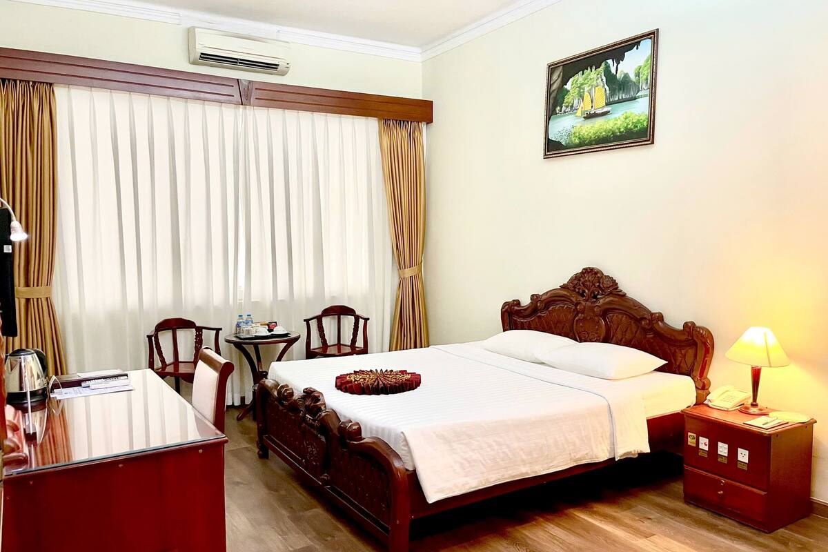 A hotel room with a wooden bed, a desk, a chair, a small table, and a lamp.