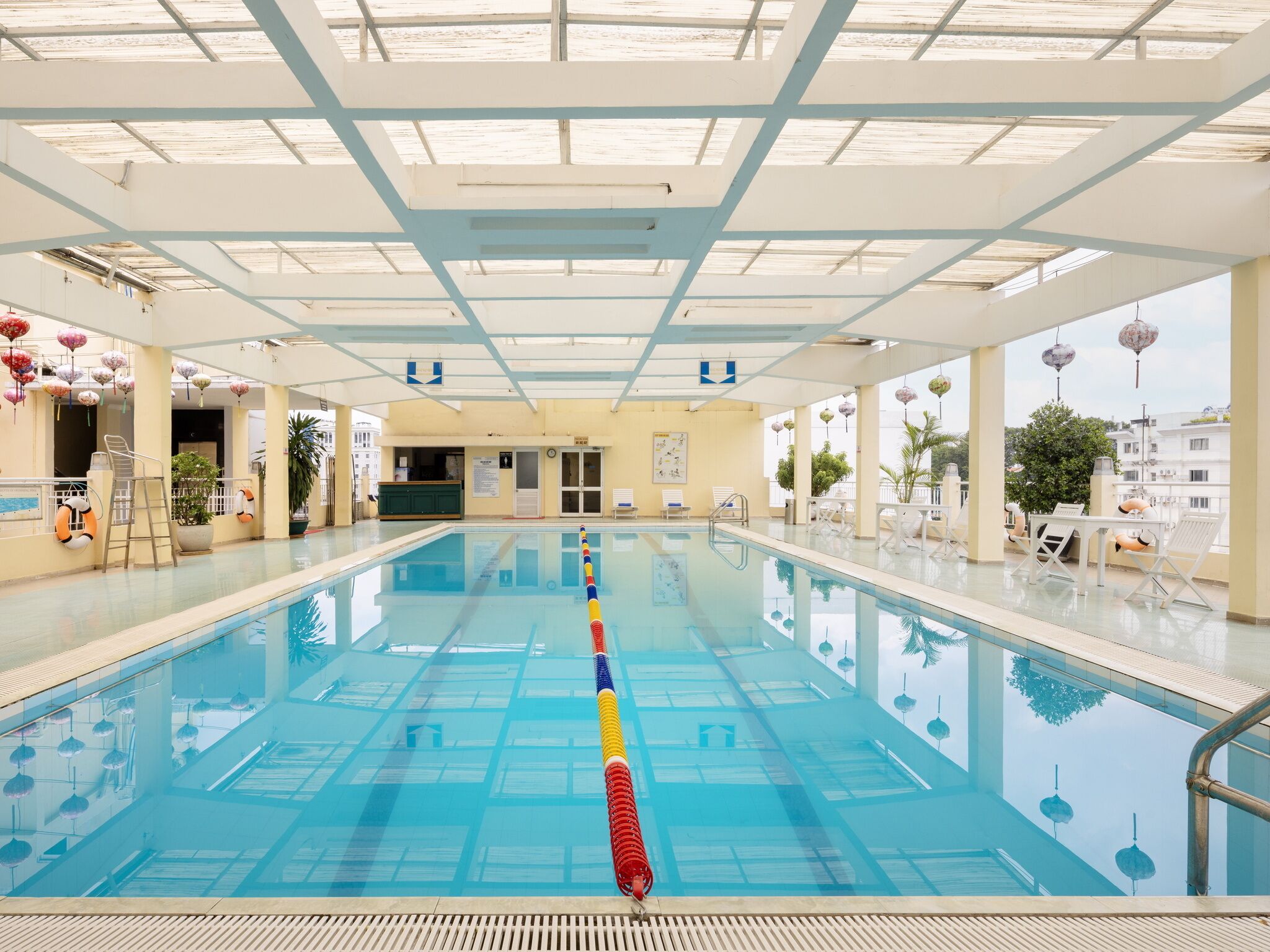 Ein Hallenbad mit deutlich markierten Bahnen, einem Rettungsschwimmerstuhl und Blick auf die Stadt durch große Fenster.