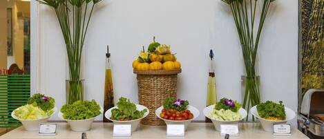 A buffet with various salads and condiments, including a bowl of mixed greens, a bowl of cherry tomatoes, and a bowl of corn salad.