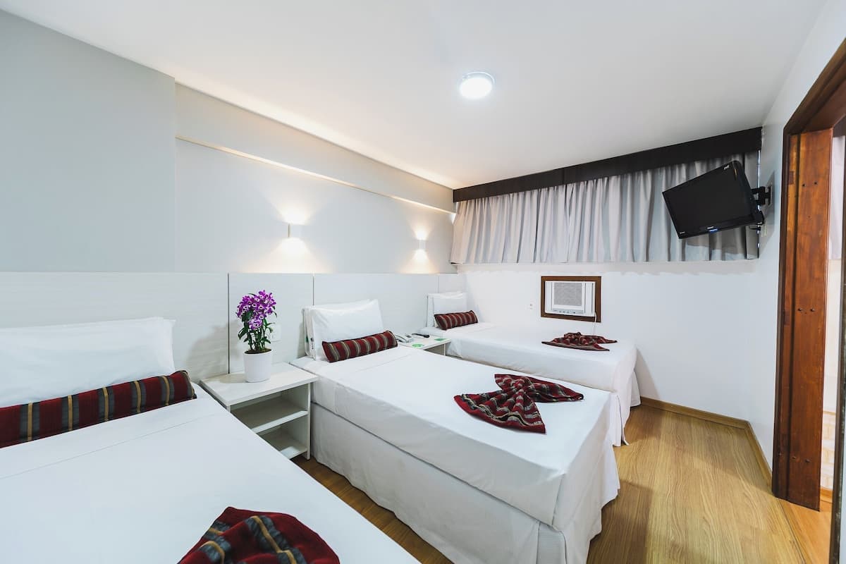 Habitación de hotel con tres camas individuales, una mesita de noche con lámpara, una planta en maceta y un televisor de pantalla plana montado en la pared.