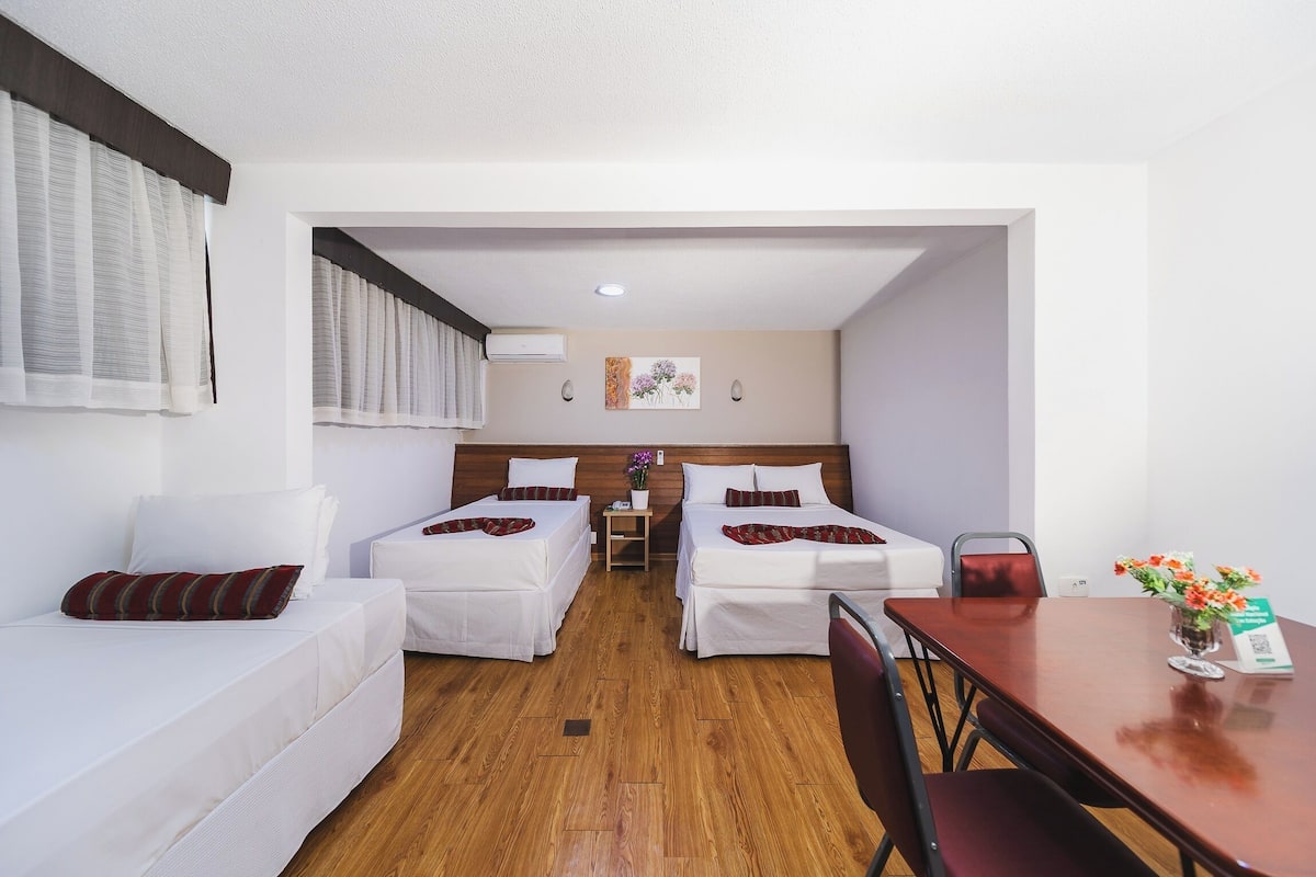 Habitación de hotel con dos camas individuales, suelo de madera, una mesa de comedor con sillas, una mesita con un florero y aire acondicionado instalado en la pared.