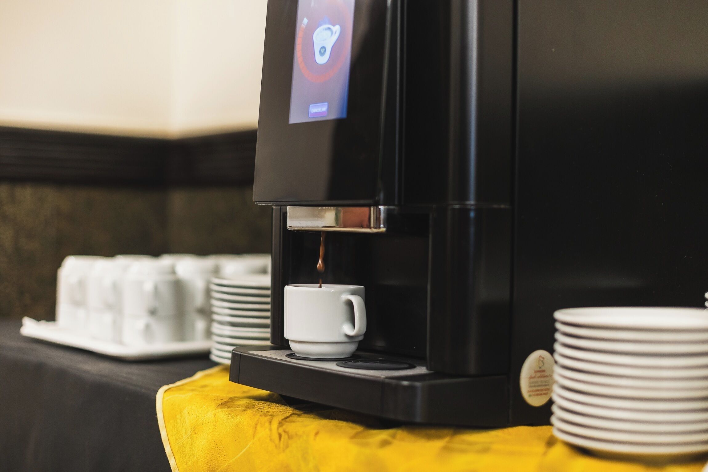 Una cafetera con pantalla digital, una taza y platos apilados sobre una mesa amarilla.