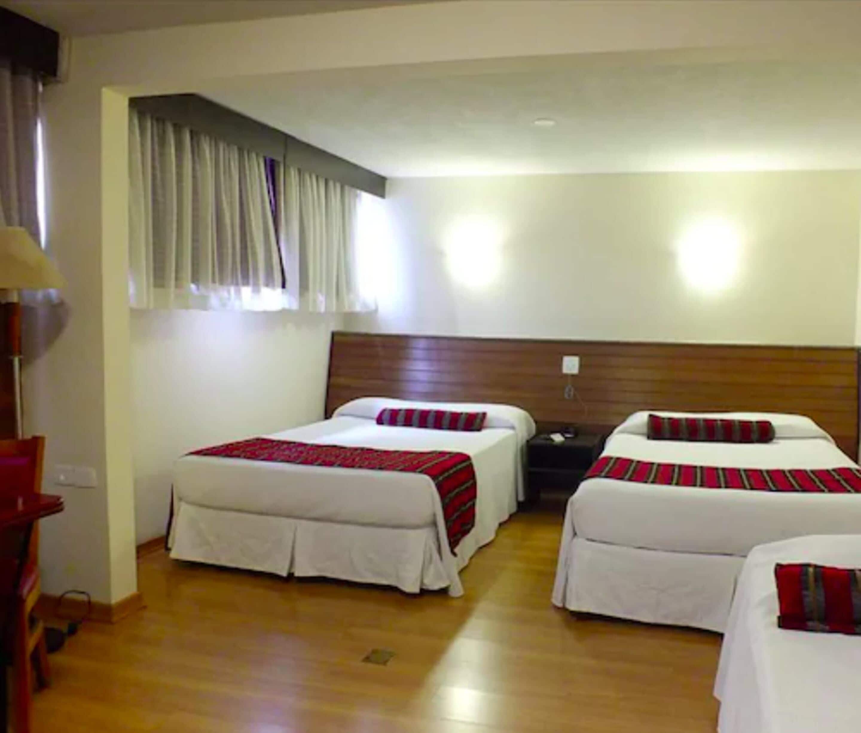 A hotel room with two single beds, a wooden headboard, a nightstand, a lamp, and a window with curtains.