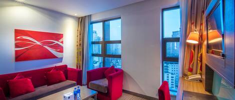 A modern hotel room with a red sofa, a small table with a remote control, and a city view through large windows.