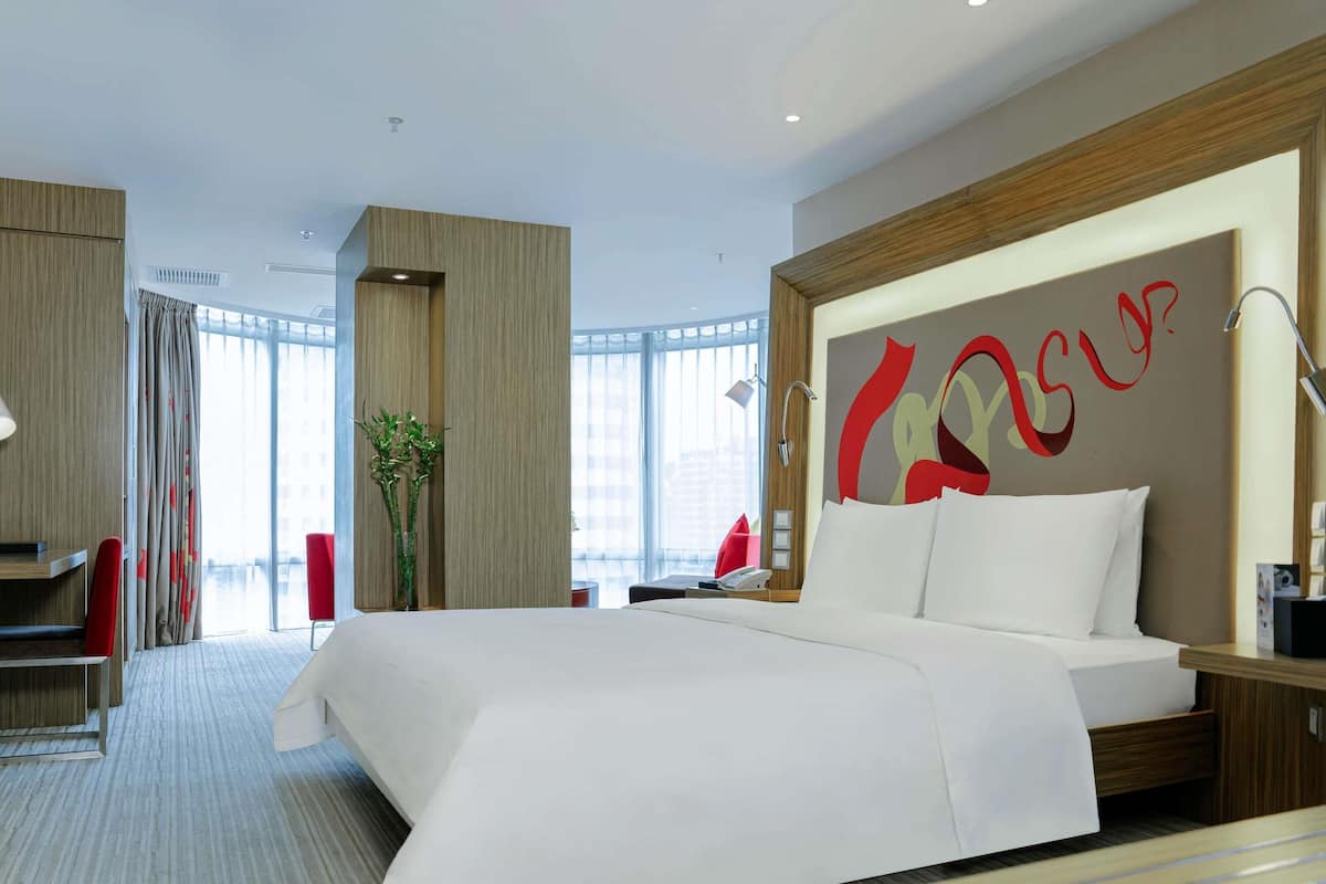 A modern hotel room with a large bed, a desk, and a chair. There is a large window with curtains, a plant, and a wall with a red and yellow design.