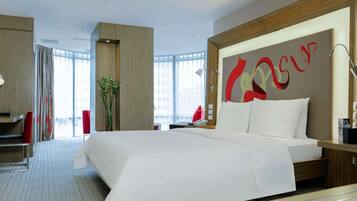 A modern hotel room with a large bed, a desk, and a chair. There is a large window with curtains, a plant, and a wall with a red and yellow design.