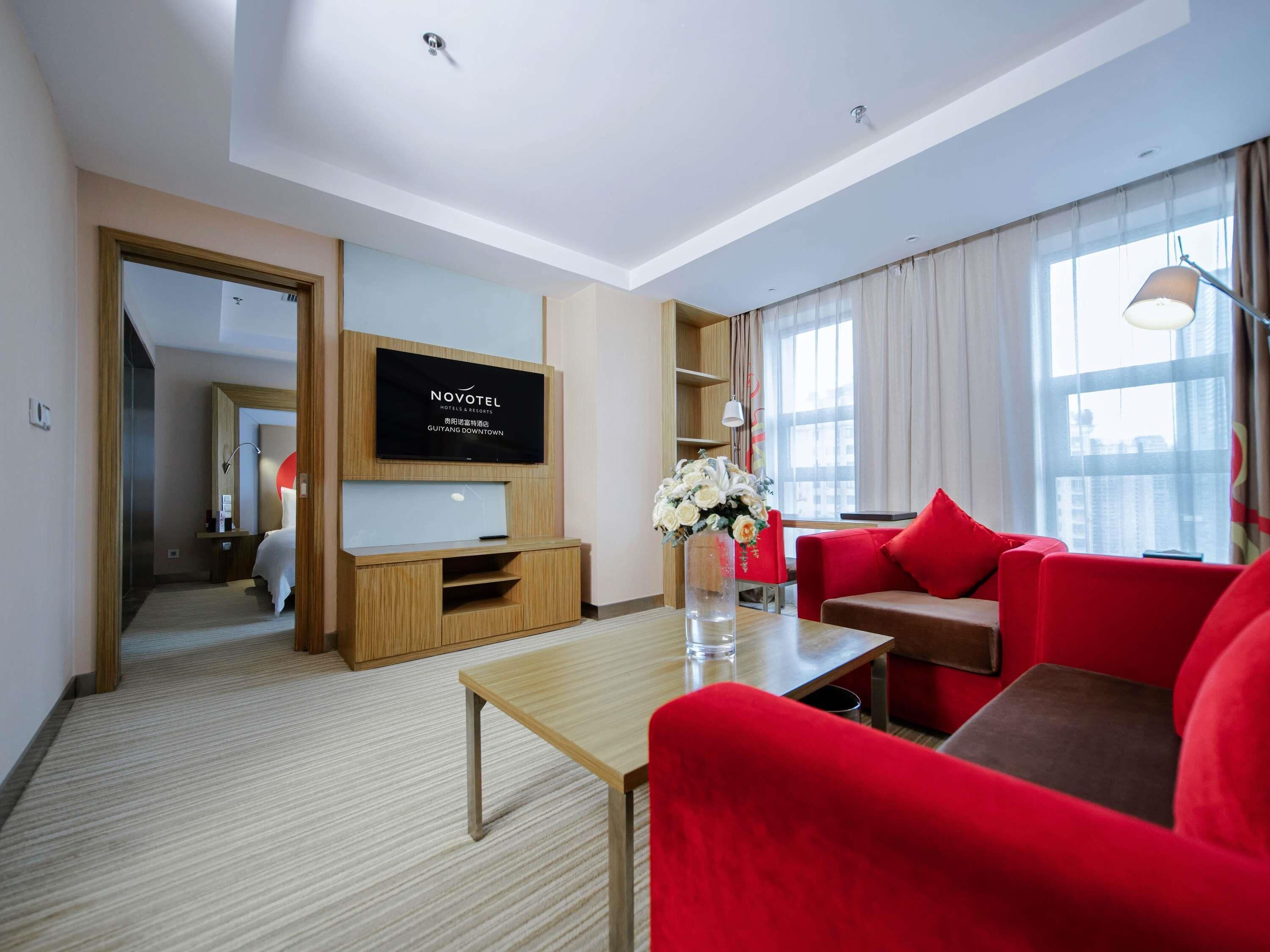A modern hotel room with a flat-screen TV, a red sofa, a wooden coffee table, and a vase with flowers.