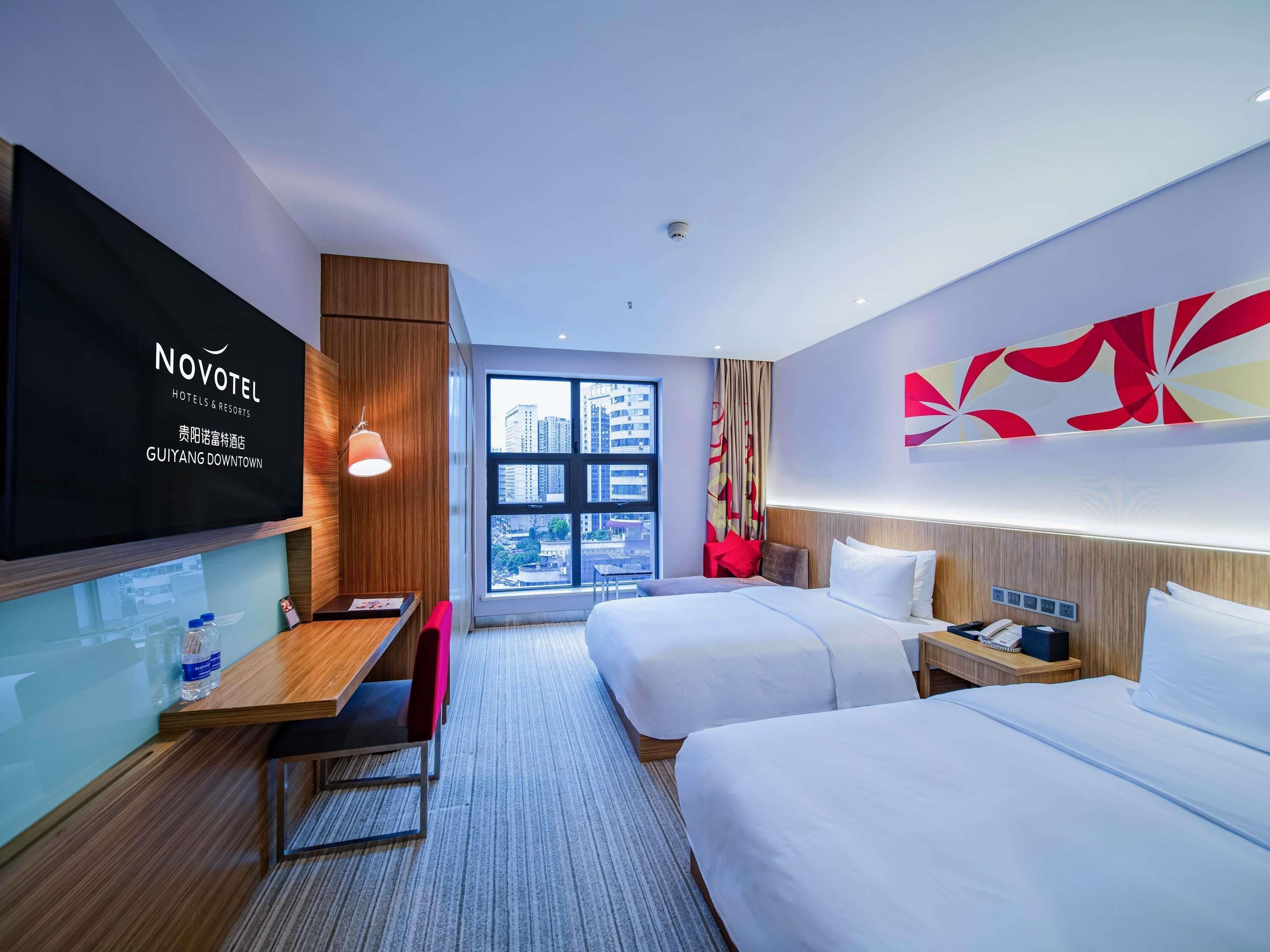 Una habitación de hotel con cama, mesita de noche y escritorio con silla. Se ve una ventana grande con vistas a edificios.