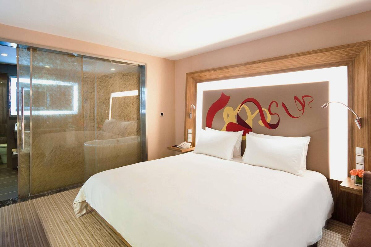 Ein Hotelzimmer mit einem großen Bett, einer Duschkabine aus Glas und einem modernen Kopfteil mit abstrakter Kunst.