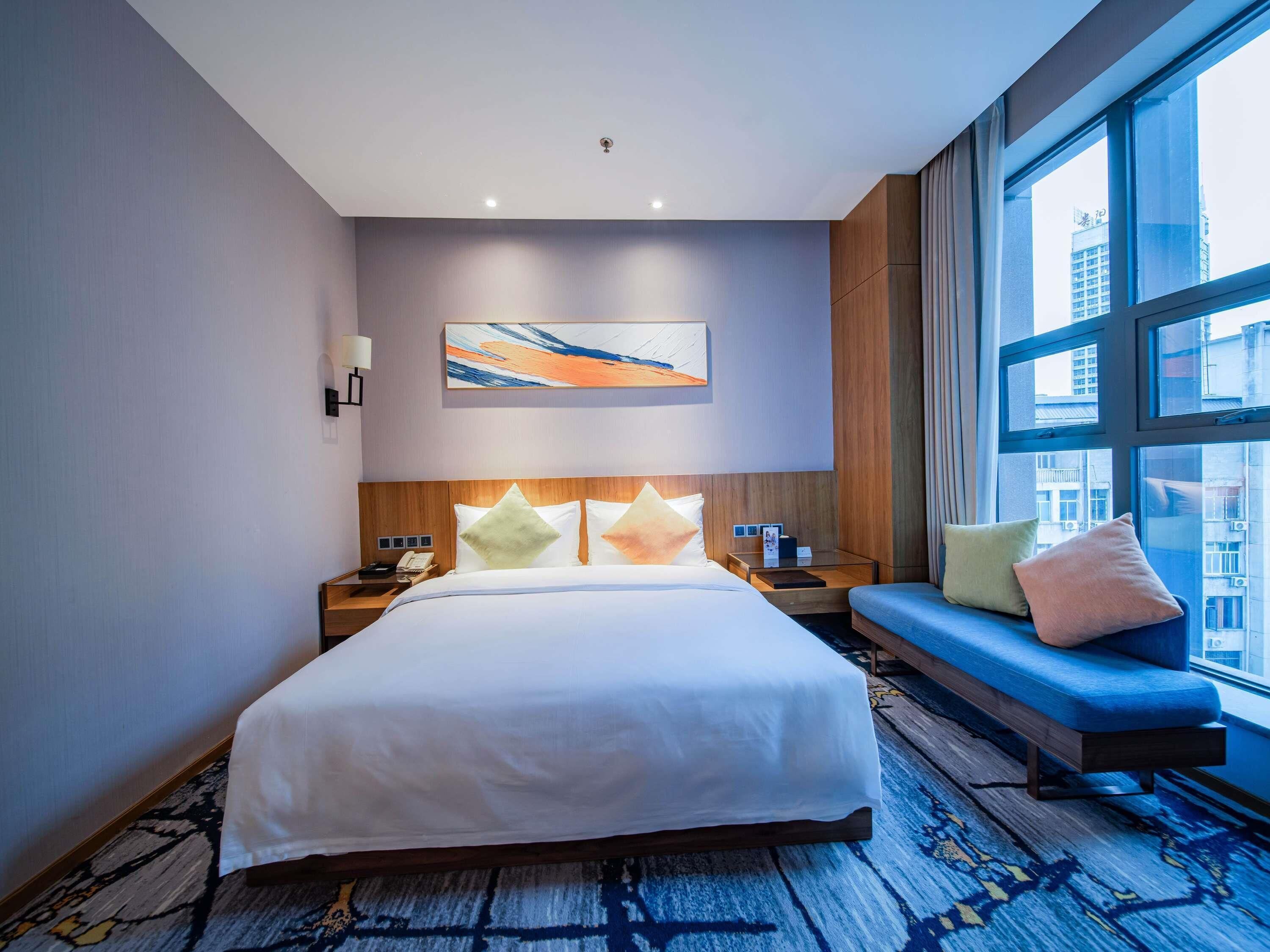 A hotel room with a large bed, a bedside table, a nightstand, a seating area with a bench, a large window, and a wall-mounted artwork.