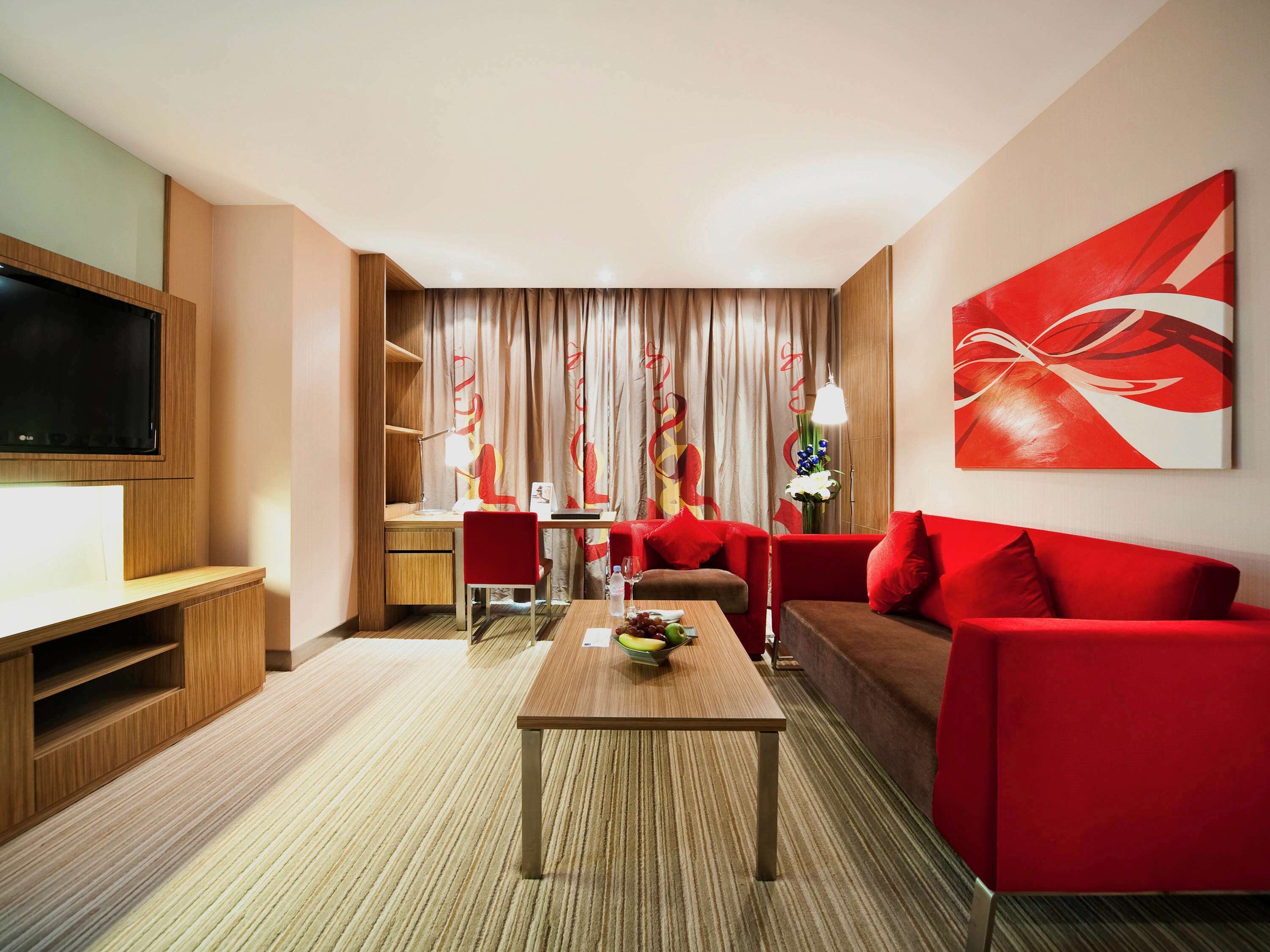 A modern hotel room with a wooden entertainment unit, a red sofa, a wooden coffee table, and a large abstract painting on the wall.