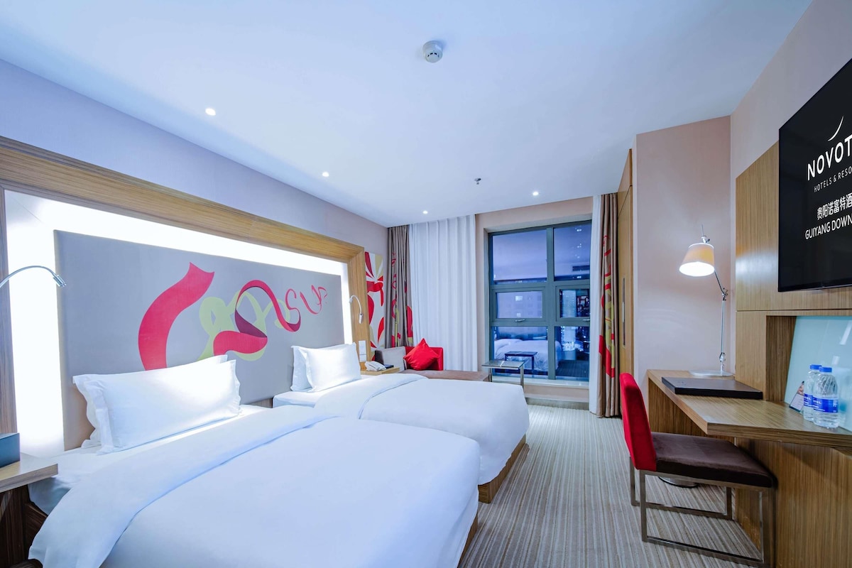 A hotel room with two beds, a desk, a chair, a TV, and a large window.