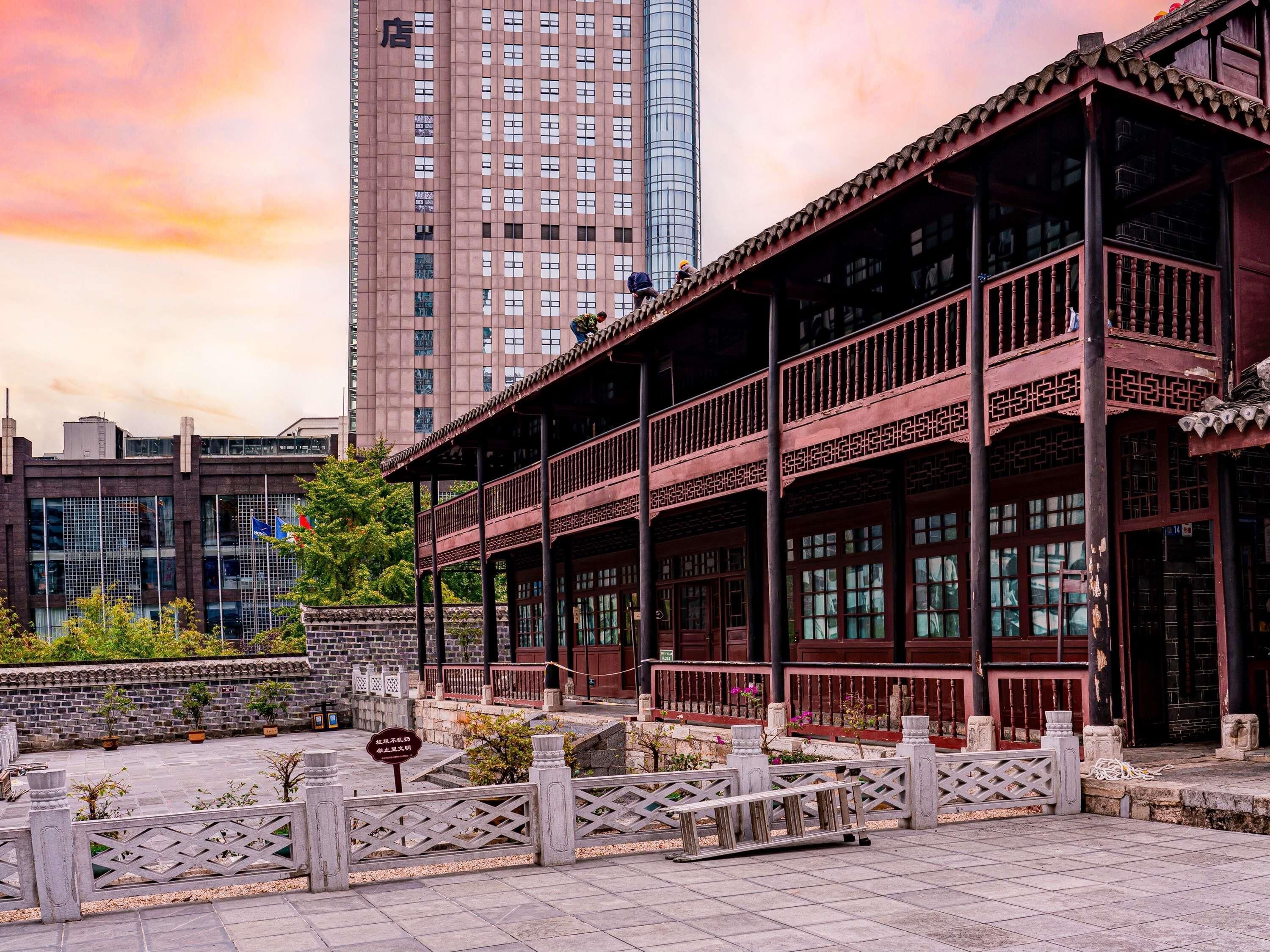 A traditional building with a balcony, a modern high-rise building in the background, and a stone wall with potted plants.