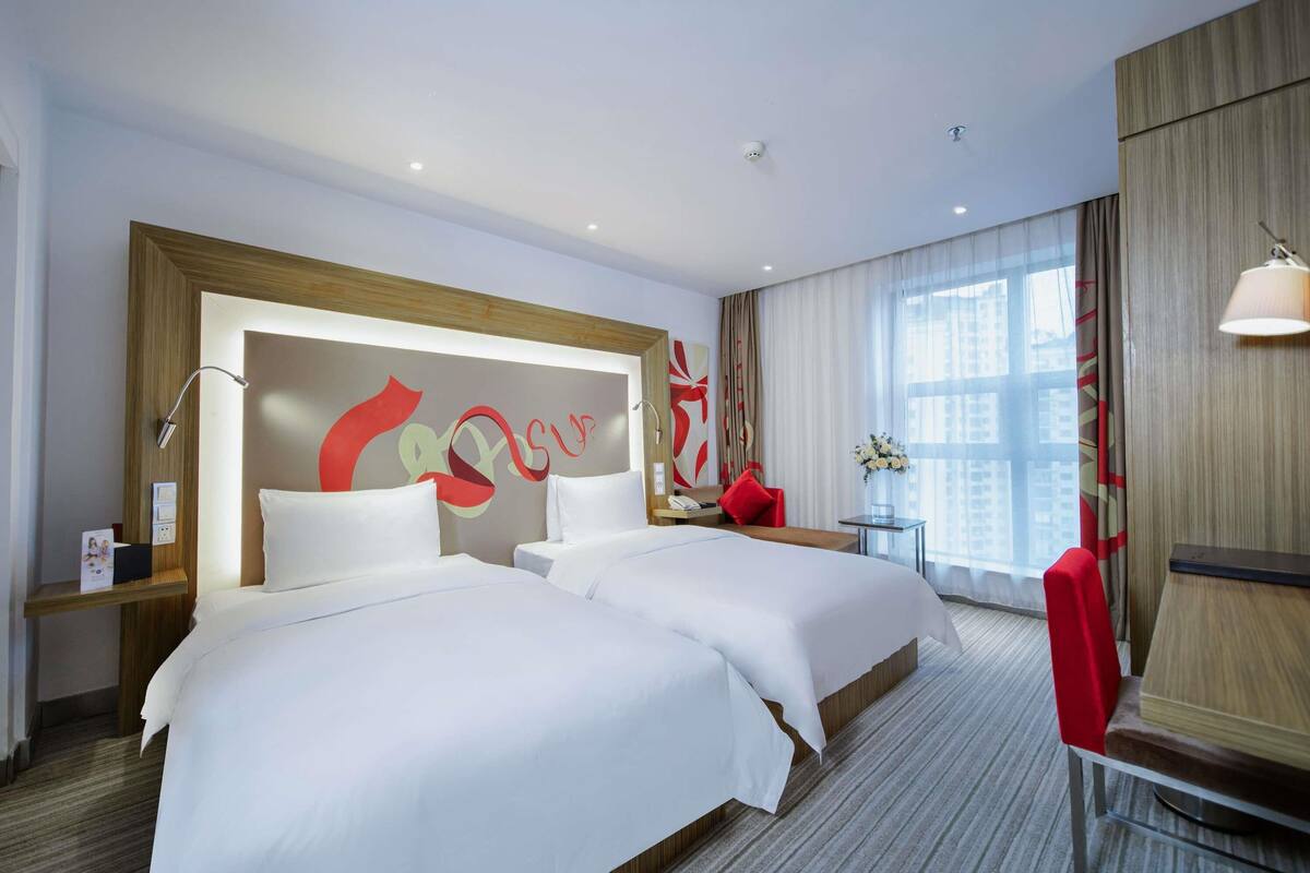 Ein Hotelzimmer mit zwei Betten, einer großen abstrakten Wandkunst, einem kleinen Tisch mit Blumen in einer Vase und einem Fenster mit Vorhängen.
