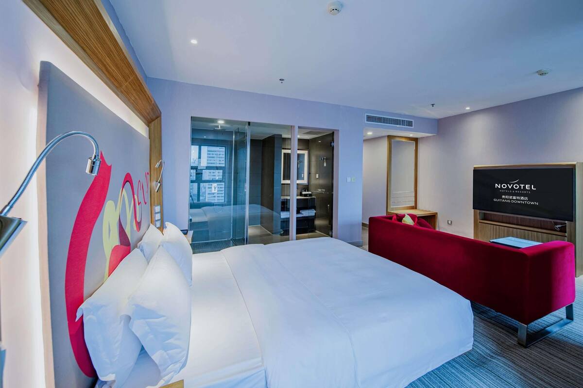 Ein Hotelzimmer mit einem Bett, einem roten Sofa, einem Fernseher und einer Glastür zu einem Badezimmer.