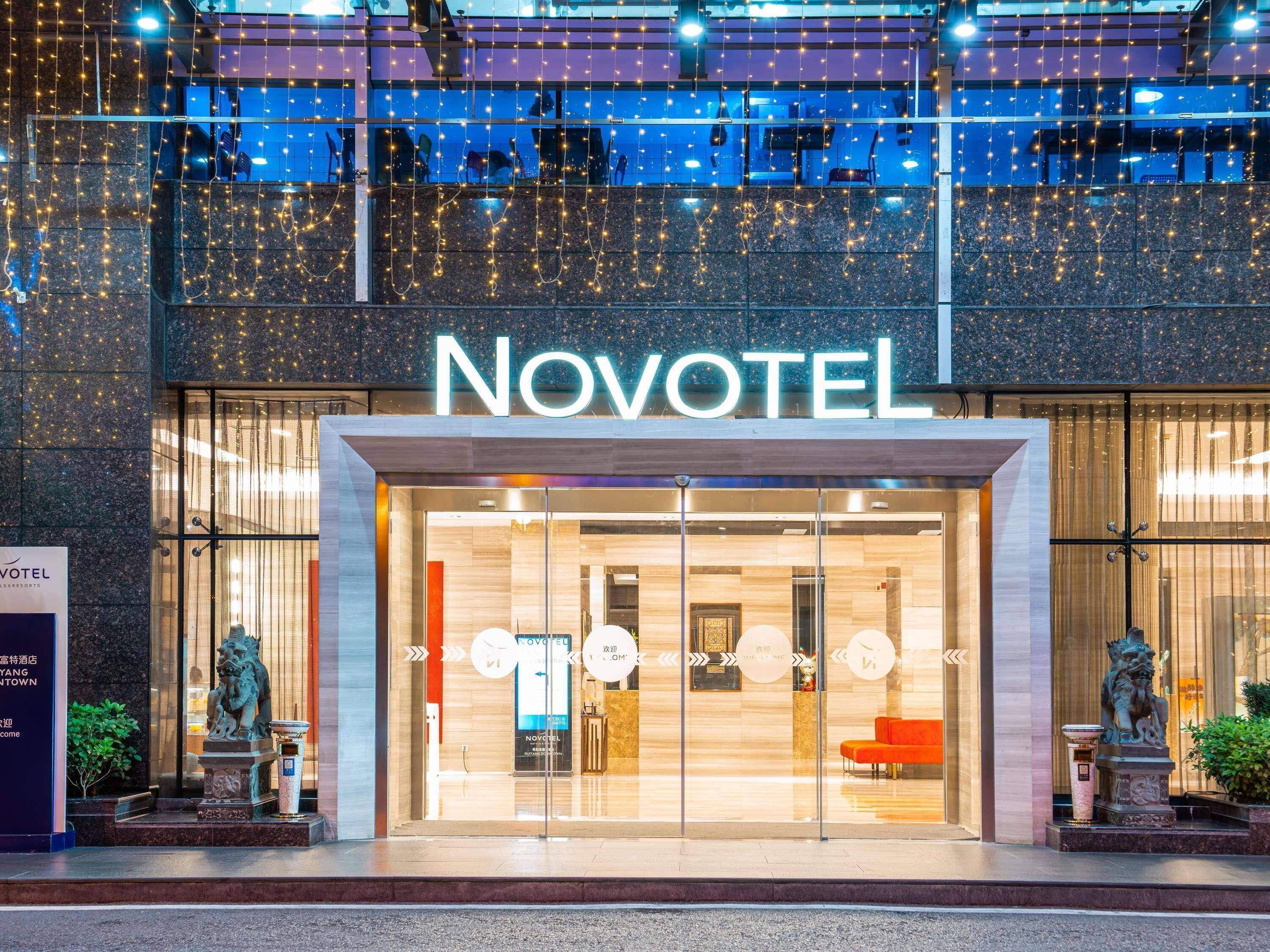 A hotel entrance with a modern design, featuring a prominent 'Novotel' sign, glass doors, and a reception area with seating.