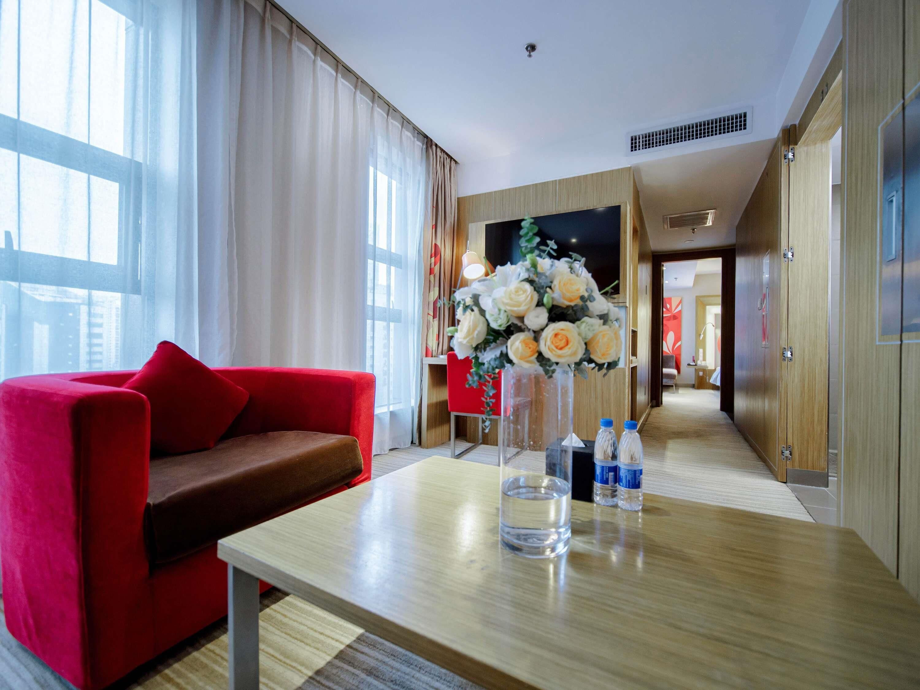Ein Hotelzimmer mit einem roten Sofa, einem Holztisch mit Wasserflaschen, einem Blumenstrauß und einem Fernseher an der Wand.