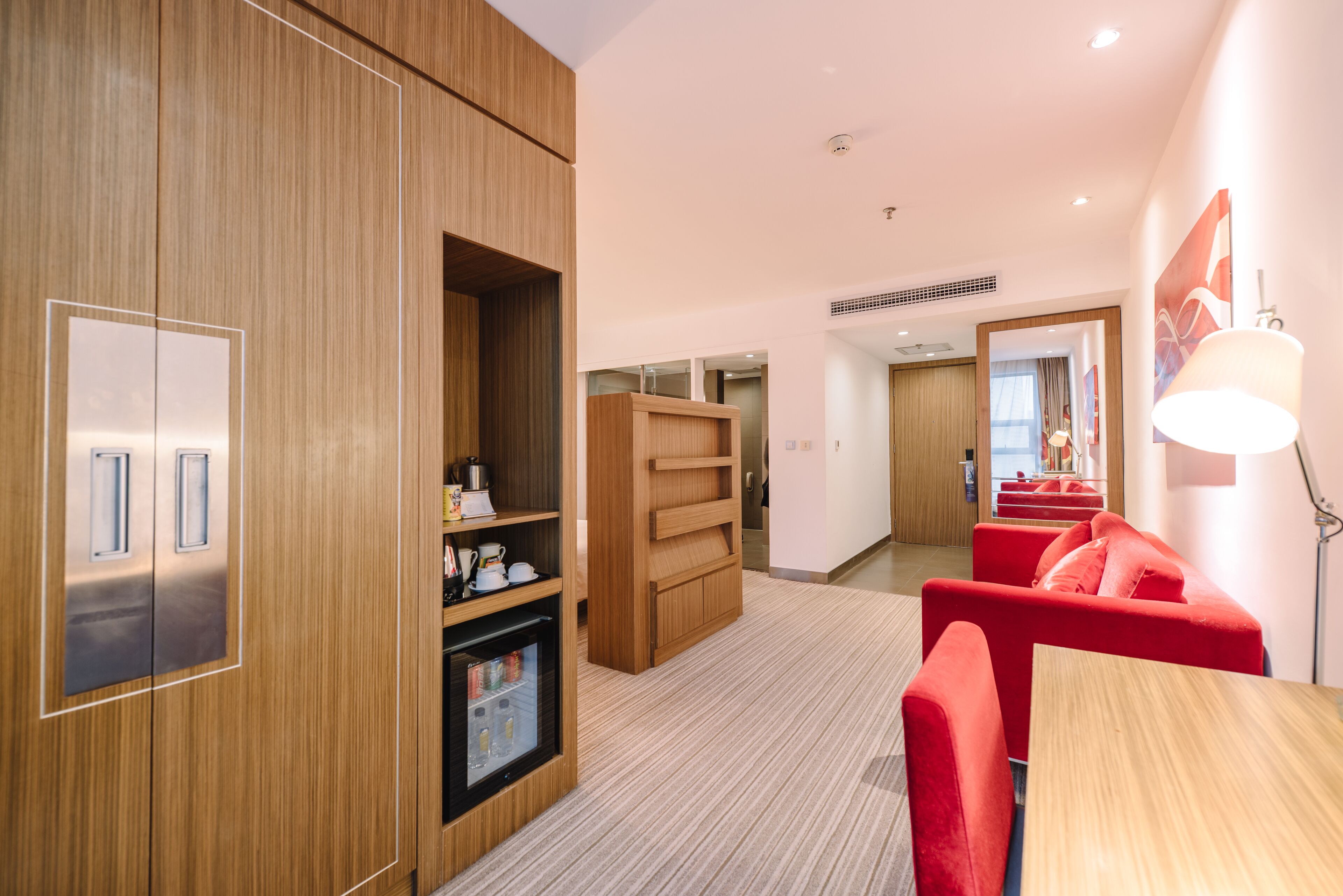 Une chambre d’hôtel moderne avec des meubles en bois, un réfrigérateur intégré, un canapé rouge et une table à manger.