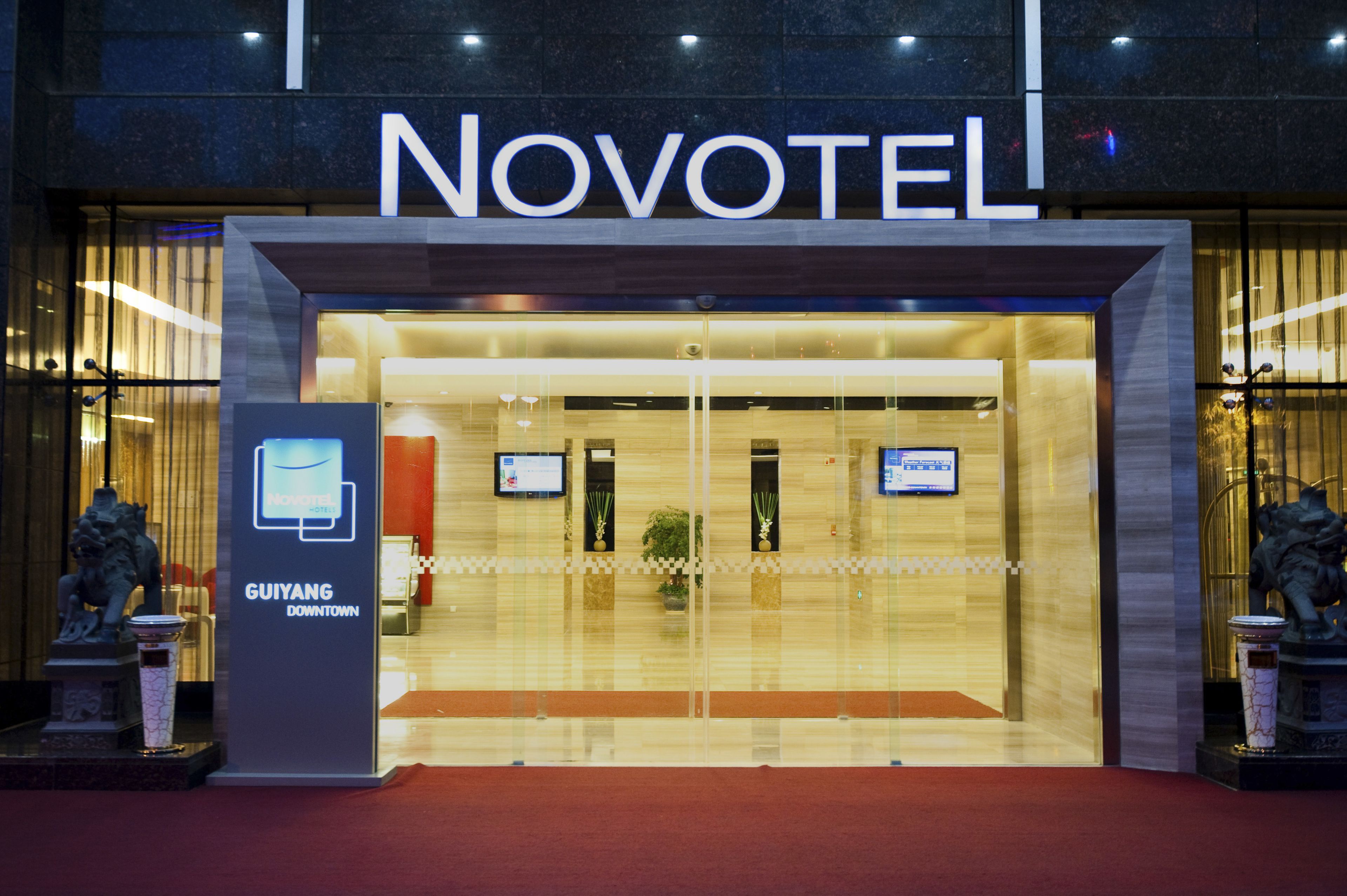 The exterior of Novotel Gyiang Downtown with a glass entrance, two statues, and a red carpet.
