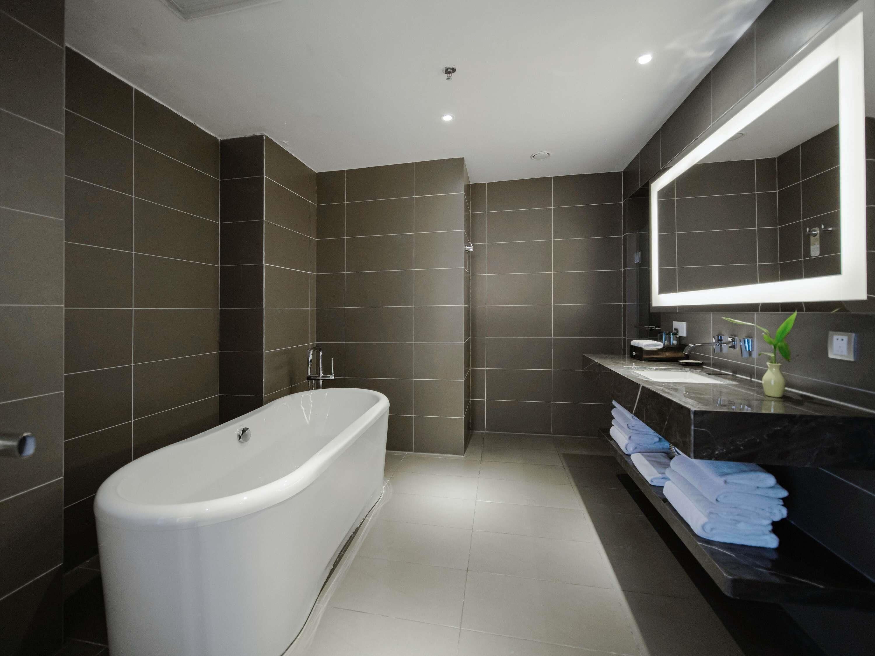 A modern bathroom with dark tiles, a freestanding bathtub, a vanity with a mirror, and neatly folded towels.