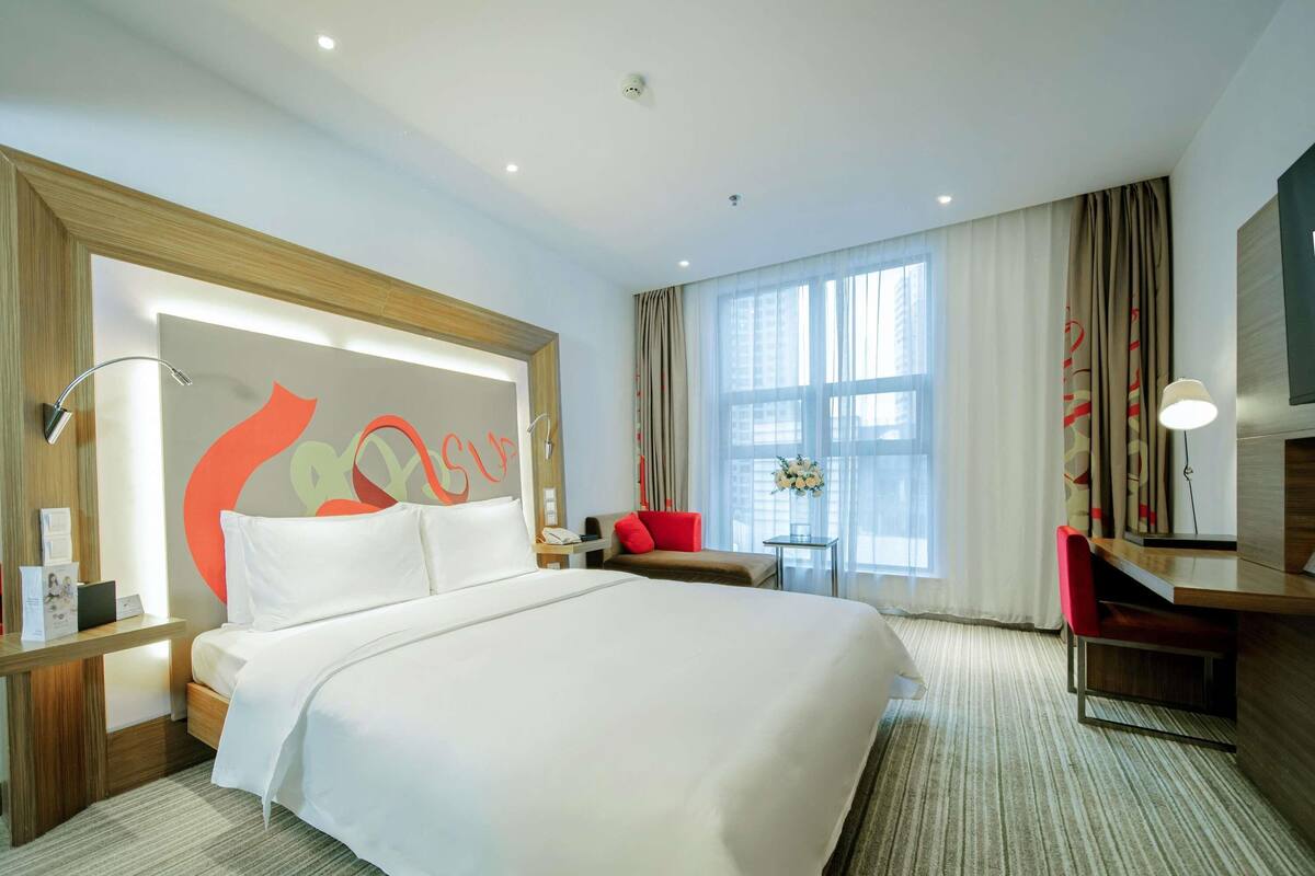Ein Hotelzimmer mit einem großen Bett, einem Schreibtisch, einem Sessel, einem kleinen Tisch mit einer Vase und einem großen Fenster mit Vorhängen.