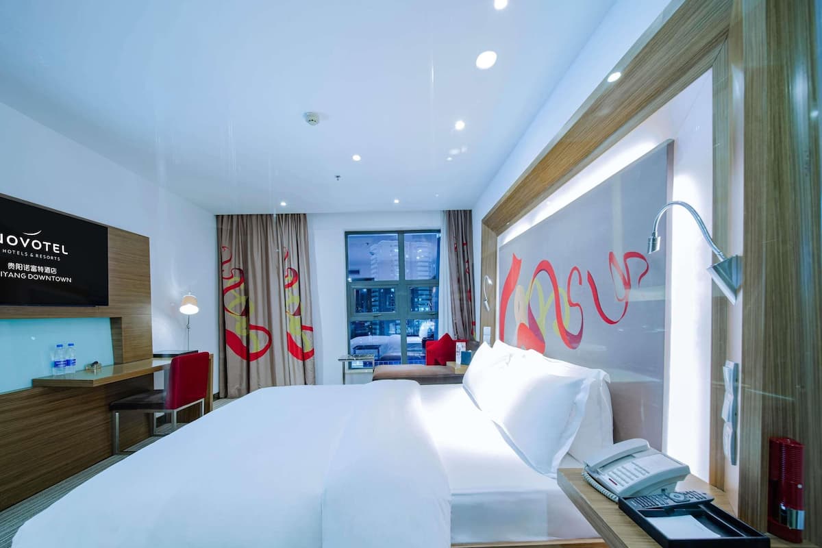 Ein modernes Hotelzimmer mit einem großen Bett, einem Schreibtisch mit Telefon und Blick auf die Stadt durch das Fenster.