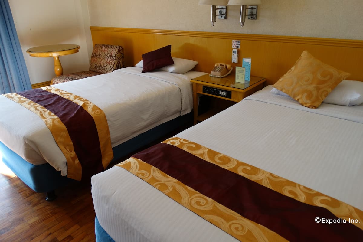 A hotel room with two beds, a bedside table with a telephone and a sign, and a small round table with a lamp.