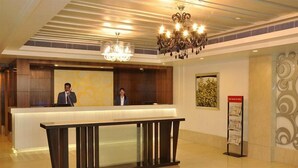 A reception area with a reception desk, two staff members, a chandelier, and a bulletin board. - Almondz Hotel (New Delhi)