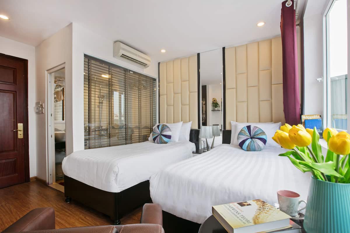 A hotel room with two beds, a large mirror, a window with blinds, a door, and a small table with a book and a mug.