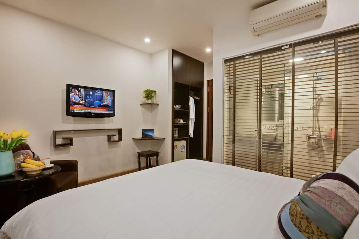 A hotel room with a bed, a TV mounted on the wall, a small table with a vase of flowers, a shelf with a plant, and a window with blinds.