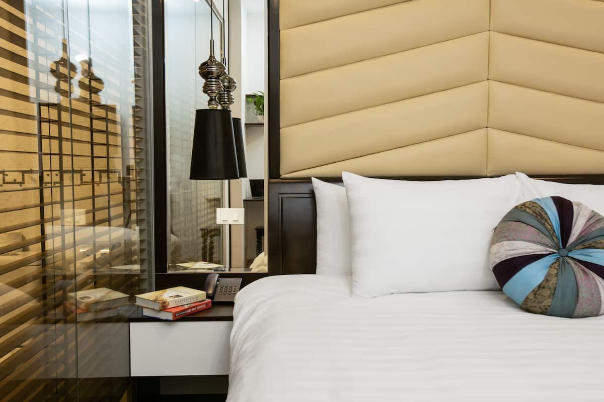 A hotel room with a modern design, featuring a large bed with white linens, a decorative pillow, a bedside table with a lamp, and a view through a window with blinds.