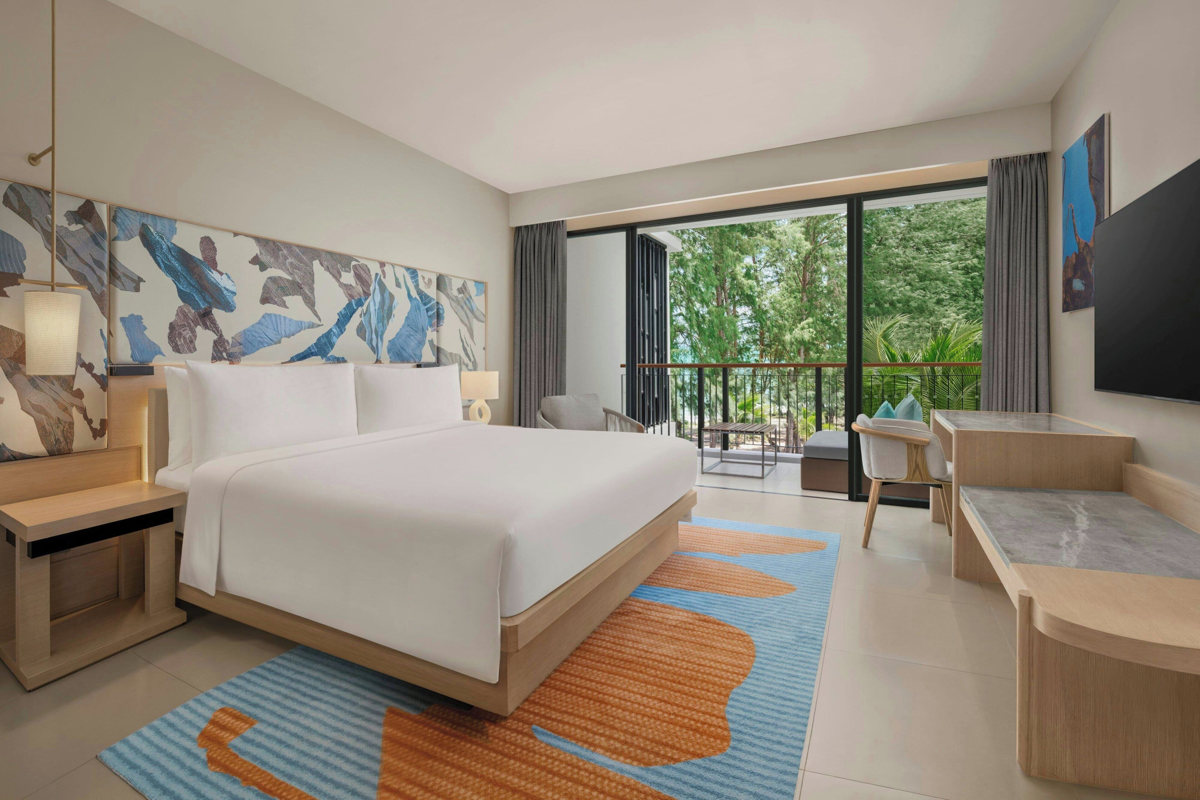 A modern hotel room with a large bed, a desk, and a balcony with a view of trees.