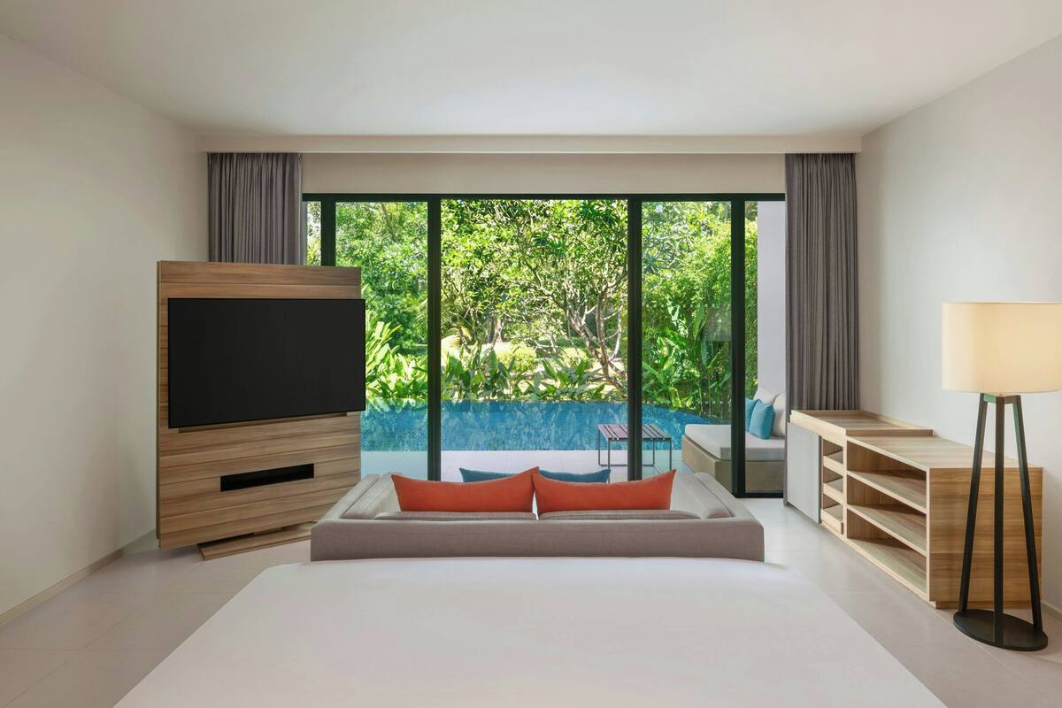 A modern bedroom with a large bed, a TV, a desk, and a view of a swimming pool and greenery.