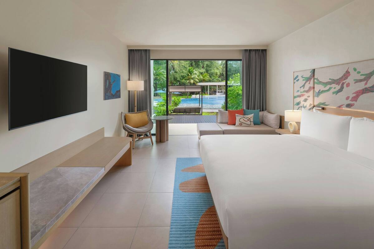 A hotel room with a large bed, a flat-screen TV, a sofa, a small table, and a view of an outdoor pool area.
