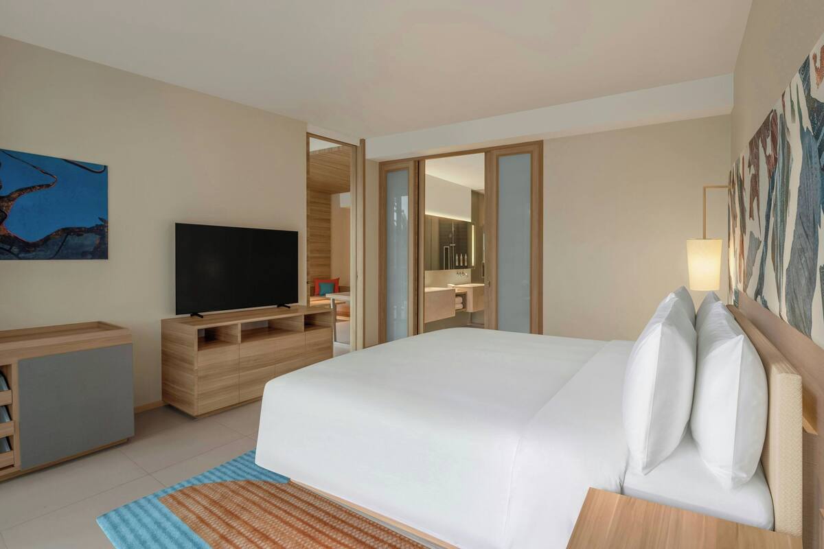 A modern hotel room with a large bed, a flat-screen TV, a wooden desk, and a bathroom visible through an open door.
